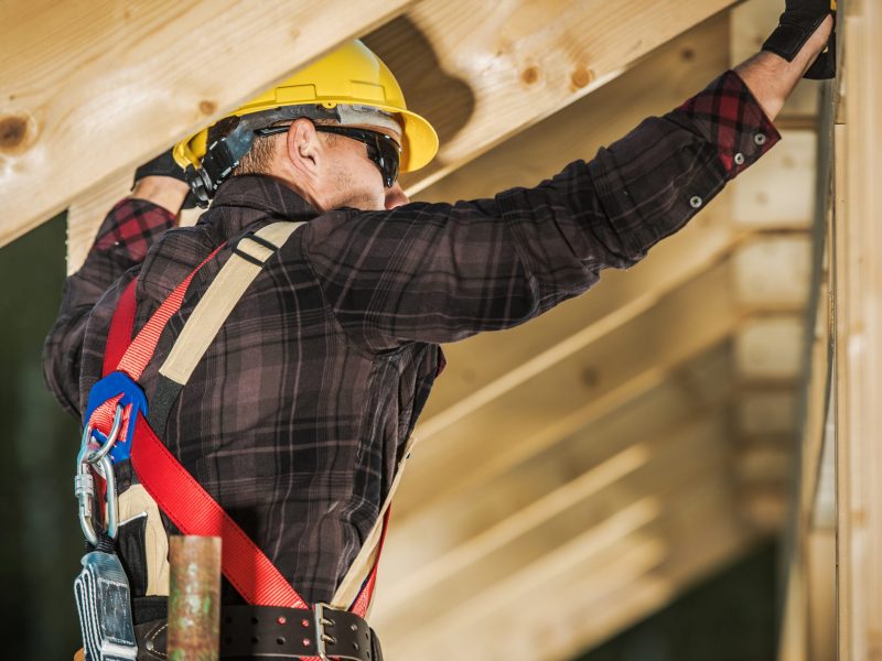A worker in a yellow helmet fixes wooden beams at a building site in the daylight.